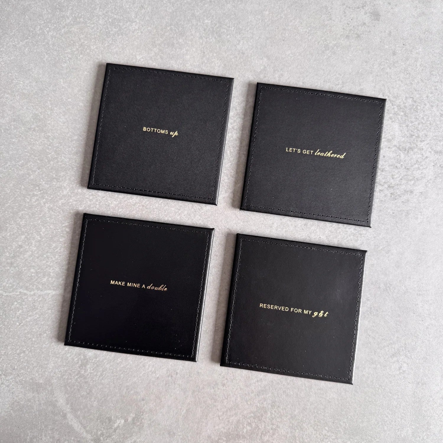 Four black leather drinks coasters in a 2x2 grid on a light gray textured surface, each with gold foil text reading different phrases including 'BOTTOMS UP', 'LET'S GET leatherbud', 'MAKE Mine a double', and 'RESERVED FOR MY GIFT'.