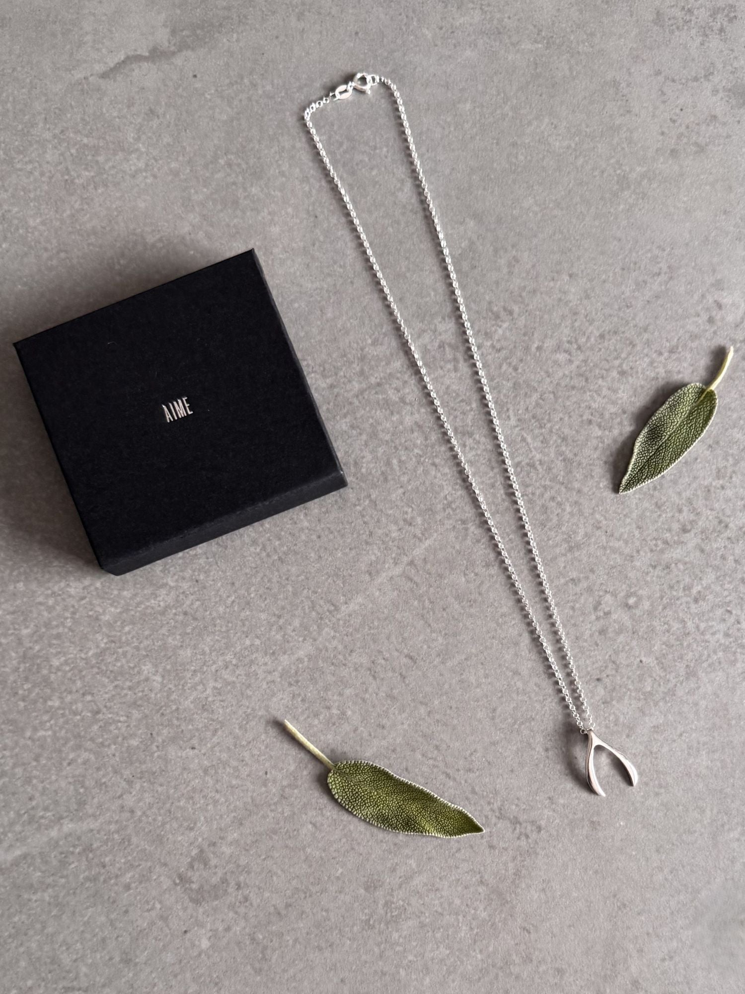 Sterling silver wishbone necklace beside a closed black Wue gift box, styled with fresh sage leaves.