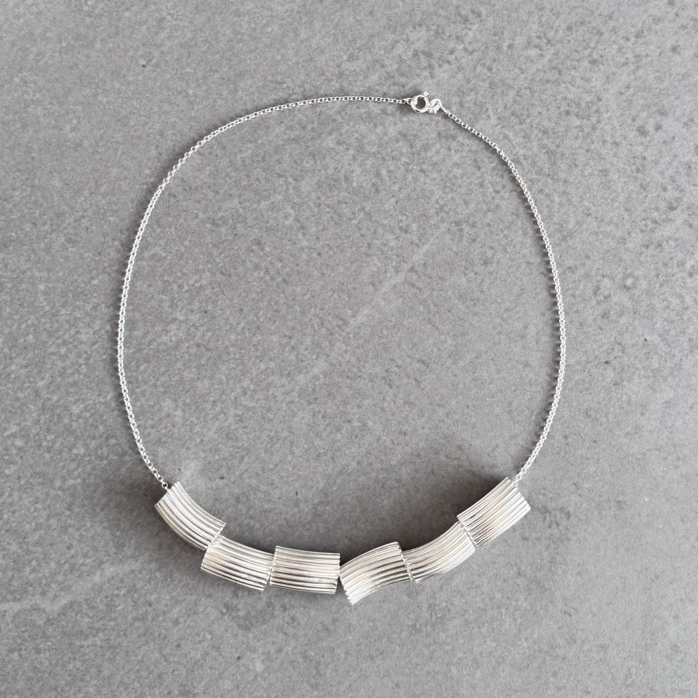 A silver necklace featuring a series of textured, ribbed rectangular links arranged in a curved row, resting on a gray stone surface.