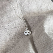 A silver pumpkin pendant necklace with a carved jack-o'-lantern face, hanging from a delicate silver chain against a textured beige linen background.