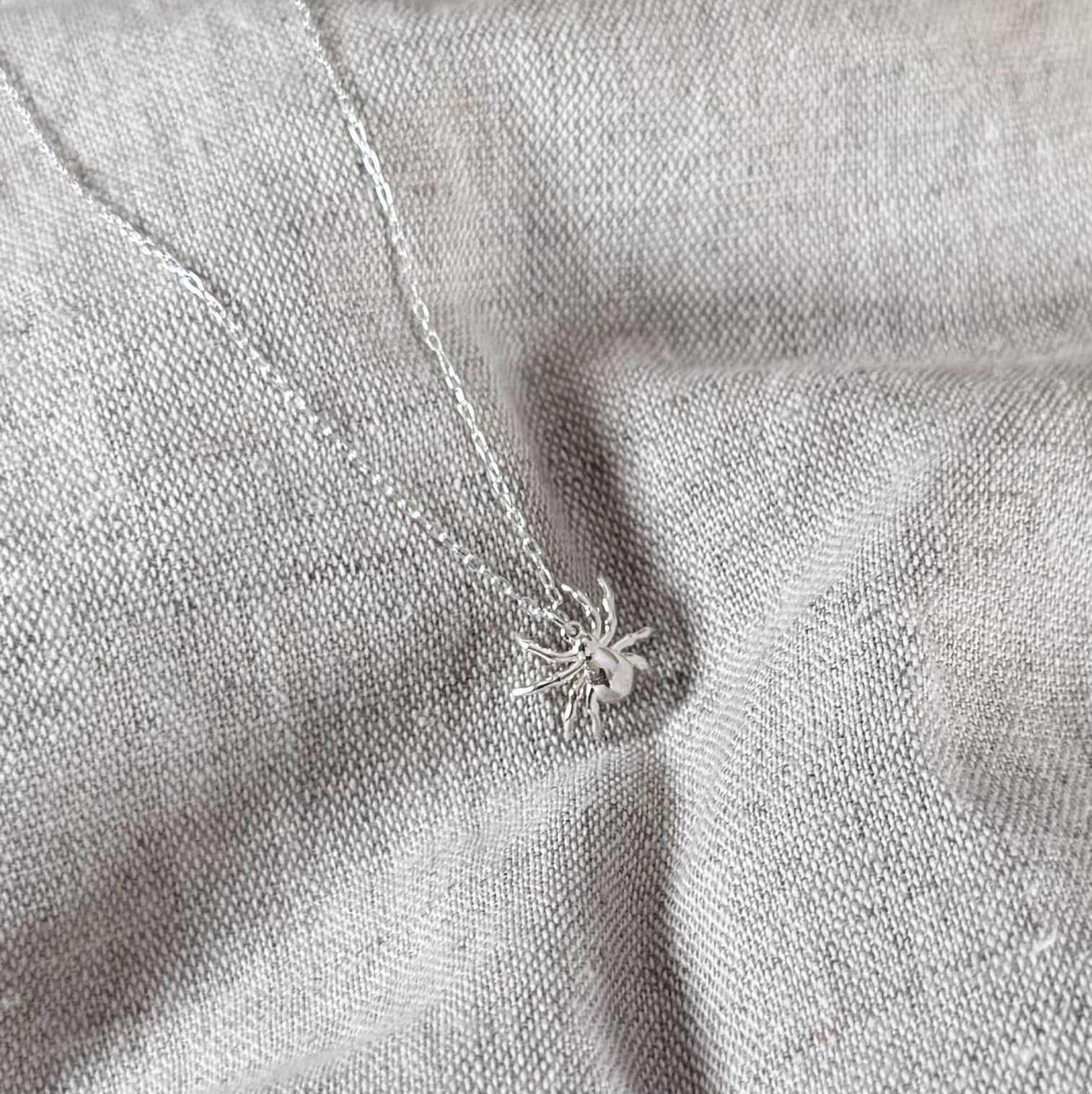A silver spider pendant necklace on a matching delicate chain, displayed on a textured light gray linen fabric background.