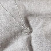 A silver spider pendant necklace on a matching delicate chain, displayed on a textured light gray linen fabric background.