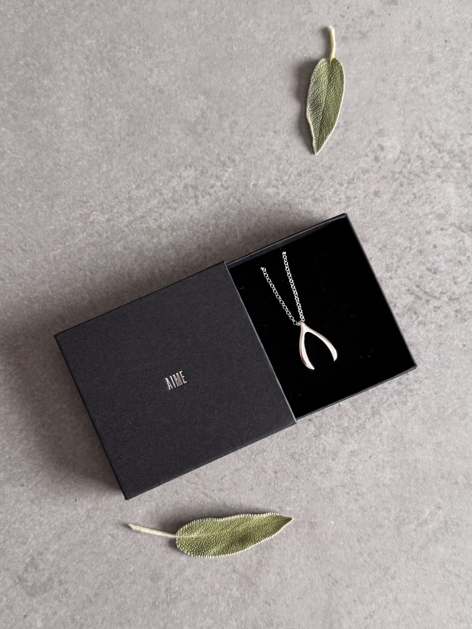 Sterling silver wishbone necklace in an open black Wue gift box with sage leaves, ready to give.