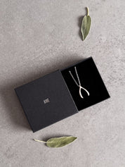 Sterling silver wishbone necklace in an open black Wue gift box with sage leaves, ready to give.