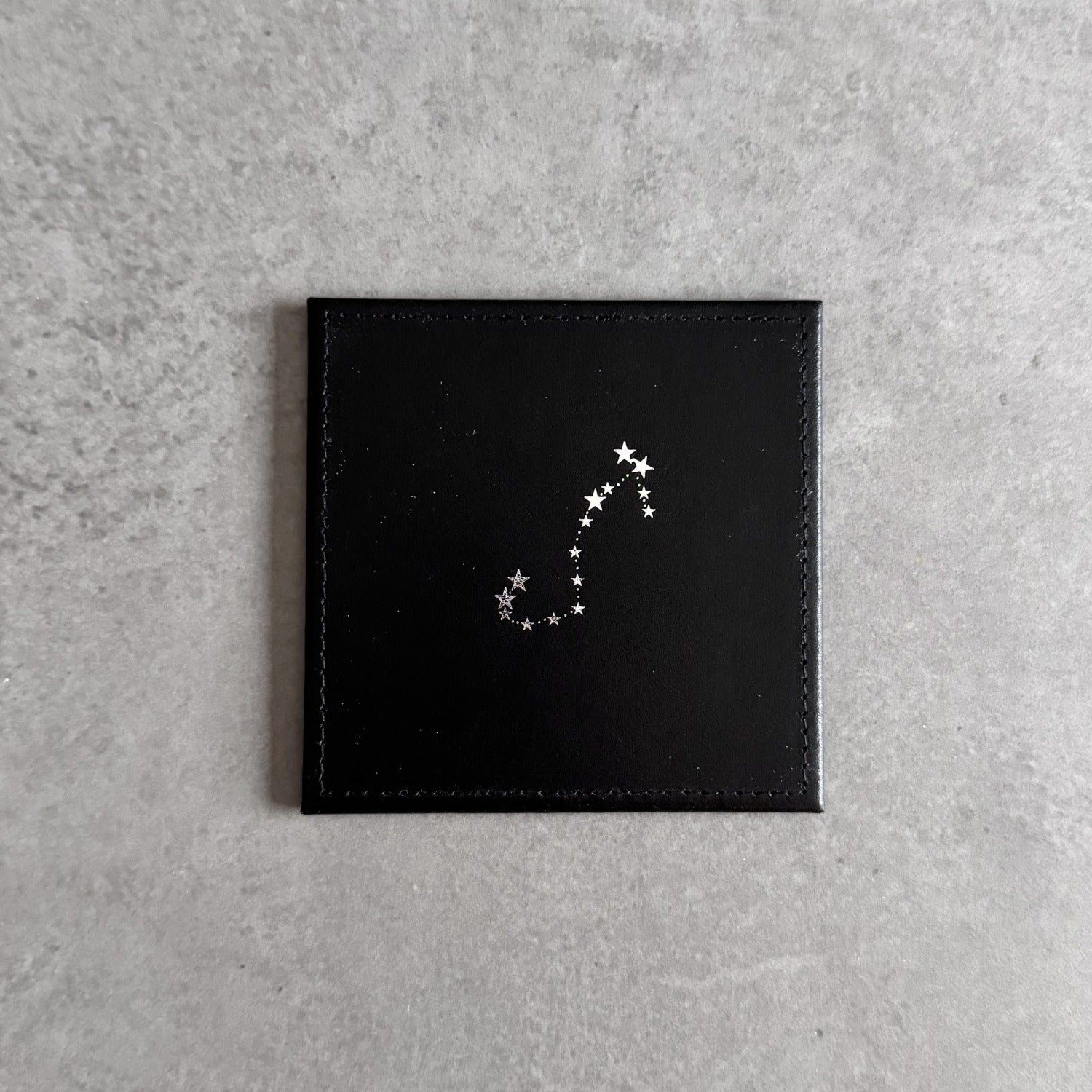 A square black leather or faux-leather card holder or wallet with a silver-toned constellation design on the front, displayed on a textured gray surface.