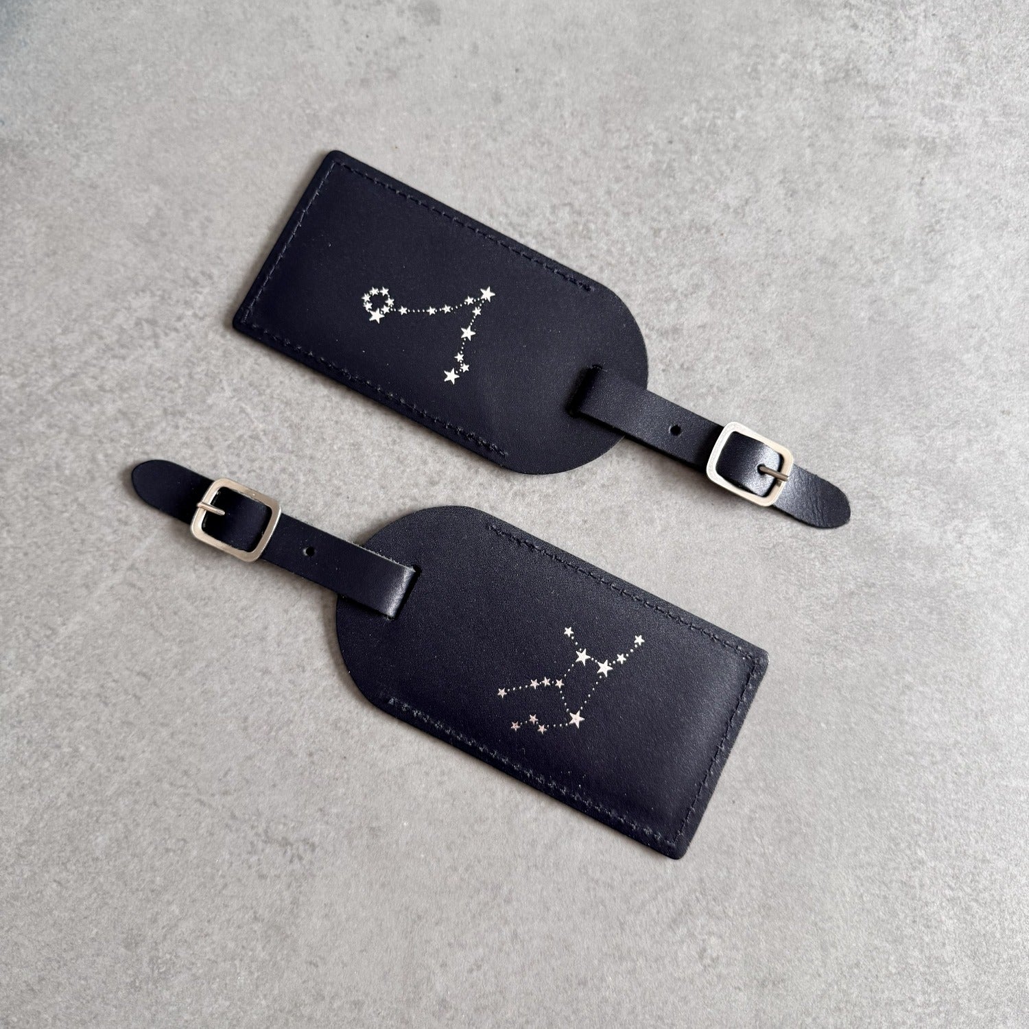 Two black leather luggage tags with silver buckle straps, featuring white constellation designs, displayed on a textured gray background.