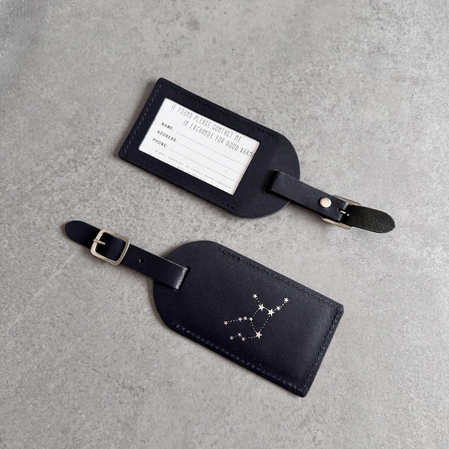 Two black leather luggage tags on a textured gray background, one with a white printed name label and the other with a silver constellation design.